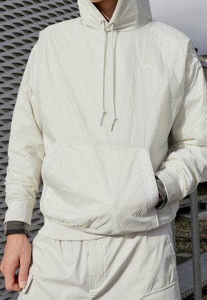 Osoba ubrana w białą kurtkę z kapturem Nike z przednią kieszenią i dopasowane białe spodnie, stojąca obok wzorzystej ściany na zewnątrz.