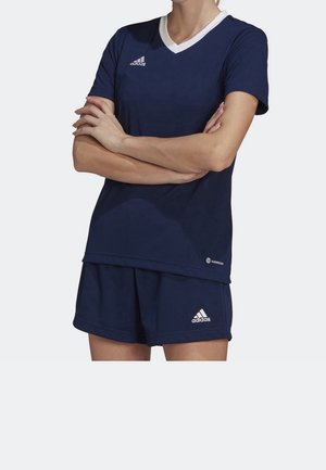Persoon gekleed in marineblauw Adidas sportkleding met witte logo's, inclusief een V-hals shirt en korte broek, armen gekruist voor de borst.