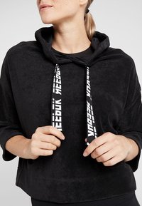 Sweat à capuche noir en tissu doux et texturé. Présente un large col, des manches longues et des cordons de serrage blancs avec logo. Coupe décontractée et confortable.