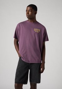 Paarse katoenen t-shirt met ronde hals, voorzien van een gele "Levi's" logo op de linkerkant van de borst. Gecombineerd met zwarte denim shorts.