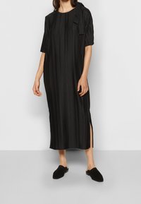 Schwarzes, plissiertes Kleid mit kurzen Ärmeln, rundem Ausschnitt, seitlichen Schlitzen und einer Schleifendetail an einer Schulter. Kombiniert mit schwarzen Mules.