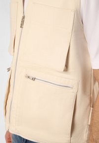 Beige Utility-Weste aus strapazierfähigem Stoff mit zwei großen Vordertaschen und einer Reißverschlusstasche in der Taille. Reißverschluss vorne.