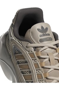 Béžová sportovní obuv s texturovaným síťovinovým svrškem, hladkými akcenty a několika hnědými pruhy. Obsahuje polstrovaný límec a tonální šněrování.