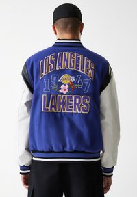 Μπλε varsity μπουφάν με κέντημα "LOS ANGELES LAKERS" σε κίτρινο, με λουλουδένιες λεπτομέρειες και λευκά δερμάτινα μανίκια. Μαύρες και άσπρες ριγωτές μανσέτες.
