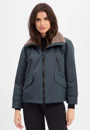 Woman with brown hair wearing a dark blue zip-up jacket with a brown fur collar and black pants against a plain light background.
