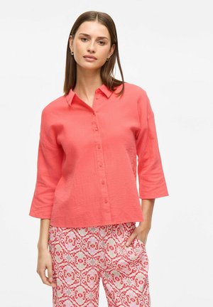 Camicia button-up color corallo con colletto e maniche a tre quarti, realizzata in tessuto testurizzato. Presenta un fit rilassato e bottoni semplici.
