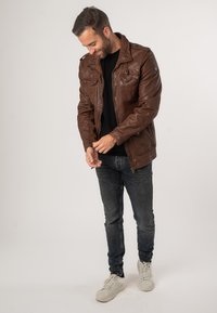 Braune Lederjacke mit Kragen, mehreren Reißverschluss- und Knopf Taschen, über einem schwarzen Shirt getragen, kombiniert mit dunklen Jeans und weißen Sneakers.
