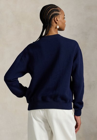 Γυναίκα με πλεξούδες, φορώντας μπλε σκούρη φούτερ και λευκό παντελόνι, στέκεται με το ένα χέρι στη μέση, γυρισμένη προς τα πίσω.