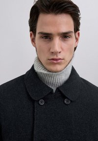 Abrigo de lana gris oscuro con cuello redondeado, abrochado con dos botones negros. El modelo lleva un suéter de cuello alto de canalé gris claro debajo.