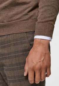 Brauner strukturierter Pullover mit gerippten Bündchen, kombiniert mit karierter Hose in Erdtönen, die ein dezentes Karomuster aufweist.