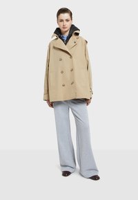 Beige kaksirivinen trench-coat puu-napeilla, suurilla sivutaskuilla ja hupulla, kerroksittain mustan hupparin ja harmaiden leveiden housujen päällä.