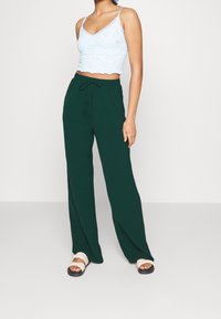 Lichtblauw kant cropped top met golvende randen, gecombineerd met donkergroene wijde broek en beige sandalen. De stoffen lijken zacht.