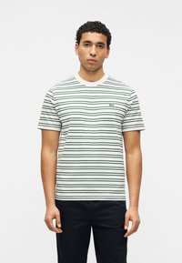 Νέος άνδρας που κοιτάζει μπροστά, φορώντας ένα κοντομάνικο μπλουζάκι με λευκές και σκούρες πράσινες ρίγες και σκούρο παντελόνι μπροστά σε απλό φόντο.