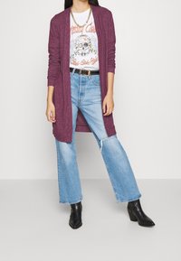 Bordeaux lång cardigan, lätt stickad, öppen front, tillsammans med en t-skjorta med grafiskt tryck med ett dödskallemönster, ljusblå slitna jeans, svarta klackboots.