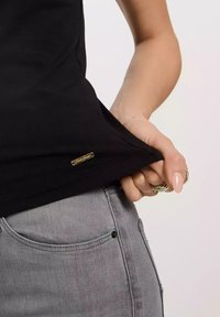 Czarny bawełniany podkoszulek z złotym metalowym metkiem przy dolnej krawędzi, w połączeniu z szarymi dżinsami z przednią kieszenią i subtelnymi detalami w szwach.