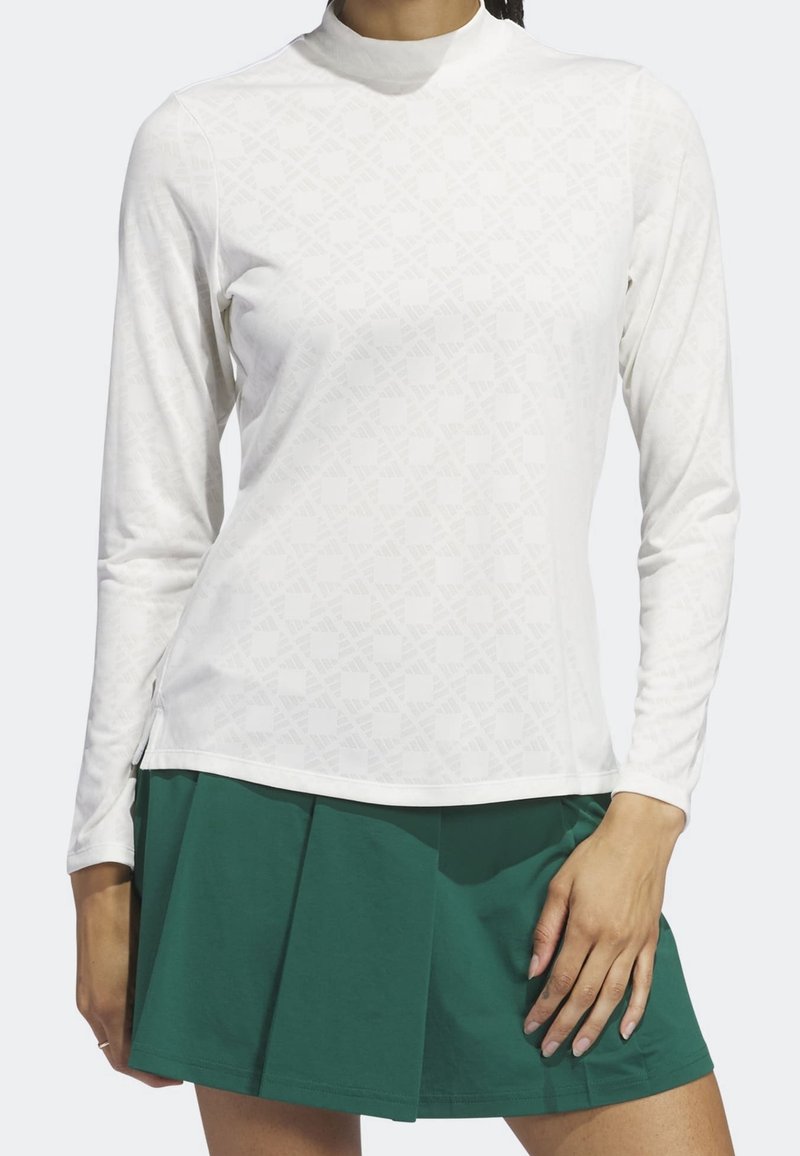 Bílé tričko s dlouhým rukávem s jemným geometrickým vzorem, s vysokým límcem a bočními rozparky, kombinované se zelenou plisovanou sukní.