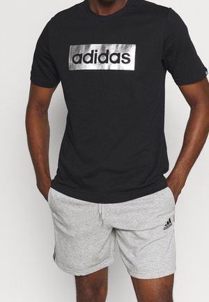 Mężczyzna w czarnej koszulce Adidas z srebrnym logo i jasnoszortach Adidas z logo na lewej nodze, ręce w kieszeniach.