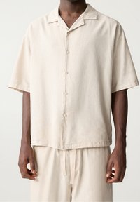 Man in een losse, korte mouw beige button-up shirt en bijpassende beige broek met trekkoord, staande tegen een effen achtergrond.