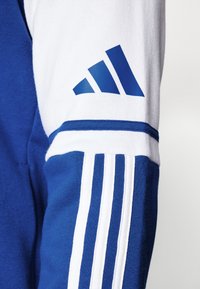 Blaues Sport Sweatshirt mit weißen Ärmeln, drei vertikalen weißen Streifen und einem blauen Logo am Ärmel. Weicher Stoff und sportliches Design.