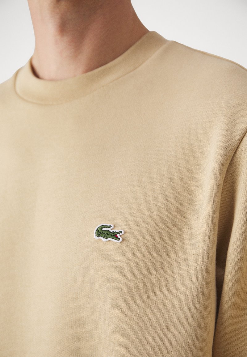 Beige Sweatshirt mit Rundhalsausschnitt, aus strukturiertem Material, mit einem kleinen grünen und roten Krokodil-Logo auf der Brust.