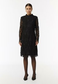 Schwarzes Spitzenkleid mit langen Ärmeln, hohem Kragen, floralem Muster, gewelltem Saum und durchsichtigen Akzenten. Tragen mit schwarzen Absätzen und Strumpfhosen.