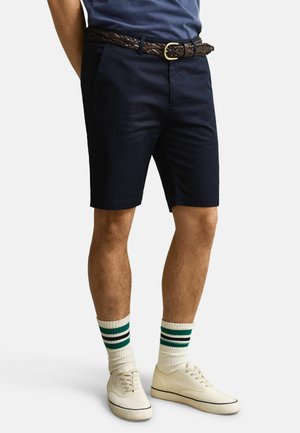 Mann iført marineblå shorts, flettet brunt belte, hvite sokker med grønne og svarte striper, og kremfargede canvas-sneakers, stående mot hvit bakgrunn.