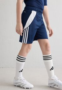 Tumši zili sporta šorti ar baltām malām, kombinēti ar baltām zeķēm ar trim melnām svītrām un baltām futbola kurpēm ar sudraba akcentiem.