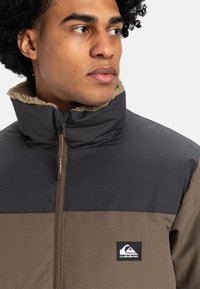 Pufferjack met een donkergrijze bovenkant en een olijfgroene onderkant, met een pluche beige kraag en een zwart logo-embleem op de borst.