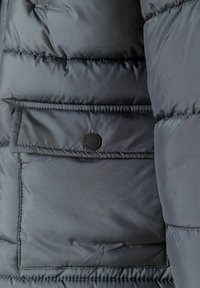 Svart puffjacka-sektion med en stor vadderad textur, en ficka med klaff och en svart cirkulär logotyp, samt slät tyg.