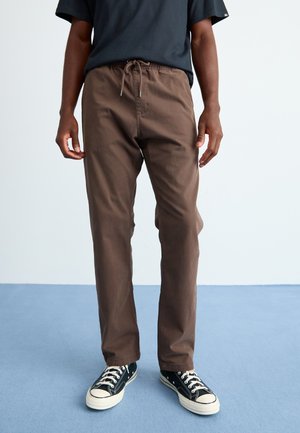 Person wearing brown drawstring pants and black-and-white sneakers, standing on blue carpet with white wall background.