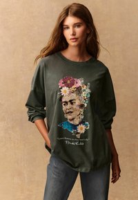 Olivenfarbener Oversize-Pullover mit einem bunten Frida Kahlo-Graphic, das Blumen und das Zitat "Ich male Blumen, damit sie nicht sterben" zeigt.