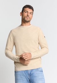 Beige langetermijn-sweater met ronde hals gemaakt van gestructureerde stof, met ribgebreide manchetten en zoom, gedragen over lichtblauwe jeans.