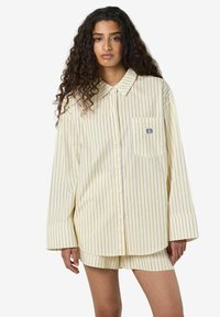 Kollase ja valge triibuline üli suur särk, millel on krae ja eesmine tasku, kooskõlas sobivate lühikeste pükstega. Kangas näib olevat kerge ja sportlik.