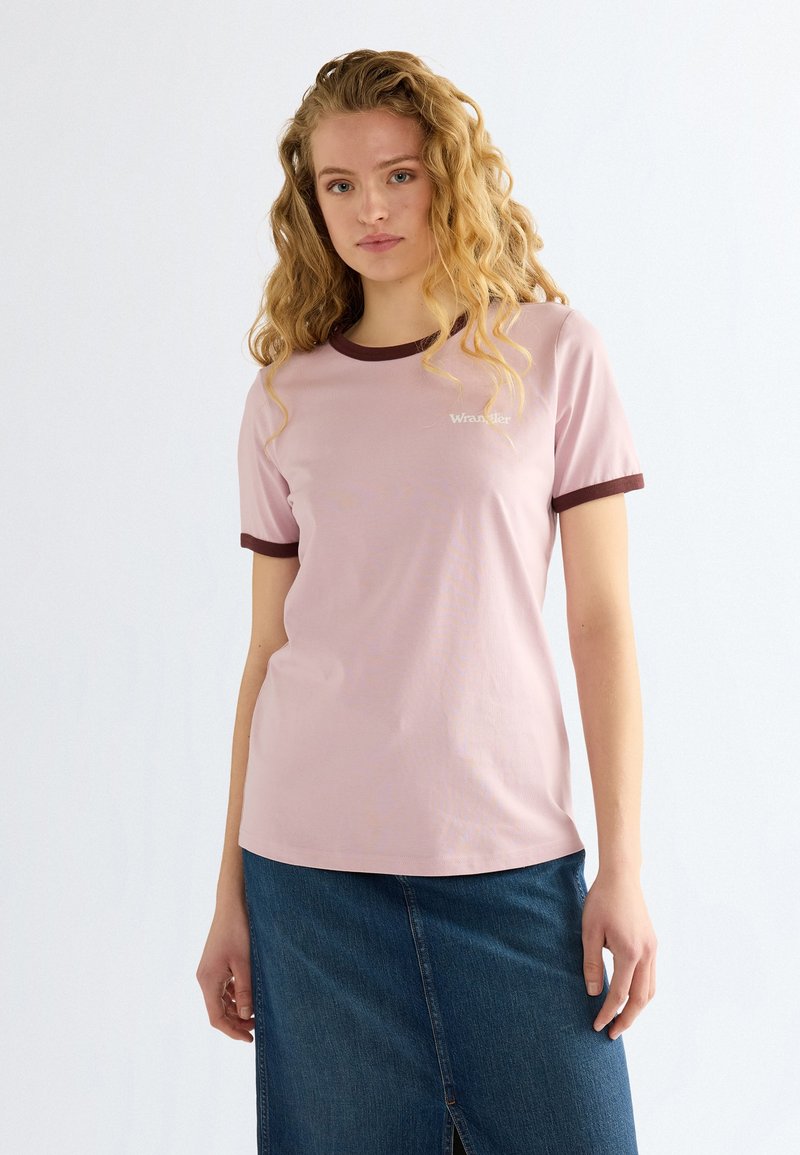 Jeune femme aux cheveux blonds bouclés portant un t-shirt rose avec une bordure marron et une jupe en jean bleue sur un fond clair uni.