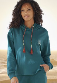 Felpa teal con maniche lunghe, cordino regolabile con perline colorate e frange. Decorata con un sole e la scritta "PARADISE".