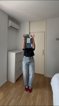 Personne portant une chemise noire, un jean bleu clair et des chaussons roses tenant un objet emballé devant son visage dans une chambre.