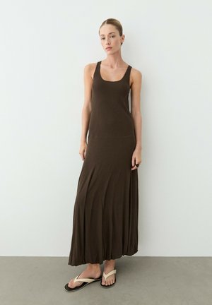 Donna che indossa un abito lungo marrone scuro senza maniche e infradito beige chiaro, in piedi contro un muro bianco semplice.