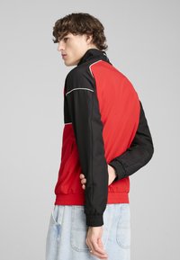 Giacca rossa e nera con colletto alto, polsini elasticizzati e tasche laterali, realizzata in un materiale liscio con colori a contrasto e accenti bianchi.
