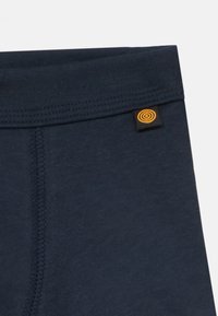 Close-up of navy blue fabric with vertical seam and small black tag featuring an orange circular logo near the waistband.