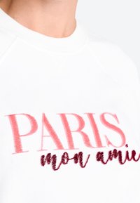 Weißer Pullover mit auffälligem, pinkem "PARIS"-Text und darunter einem kleineren, dunkelroten "mon amie" in einem weichen, strukturierten Stoff.