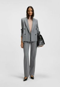 Gray double-breasted blazer with peak lapels, paired with gray tailored trousers. Model holds a black leather handbag. Smooth texture throughout.
