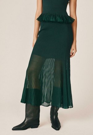 Falda midi plisada verde oscuro con sección inferior transparente, combinada con botas negras hasta la mitad de la pantorrilla con punta afilada sobre fondo neutro.