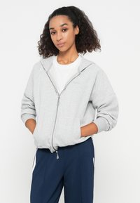 Light gray zip-up hoodie with a smooth texture, featuring a hood, kangaroo pockets, and ribbed cuffs. Paired with navy trousers.