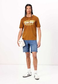 Camiseta de algodón marrón con estampado "JACK & JONES", combinada con pantalones cortos de denim azul claro. Zapatillas blancas con acentos negros, sosteniendo una toalla blanca.