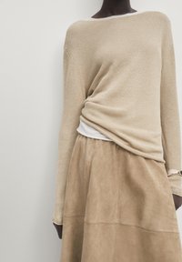 Pull beige léger en maille avec un col bateau, superposé à un top blanc, associé à une jupe beige en daim avec une texture lisse.