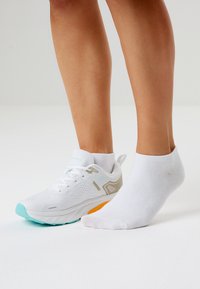 Valkoinen urheilukenkä, jossa turkoosit ja oranssit yksityiskohdat, hengittävä verkkopäällinen ja kuvioitu pohja. Valkoinen nilkkasukka paljaalla jalalla.