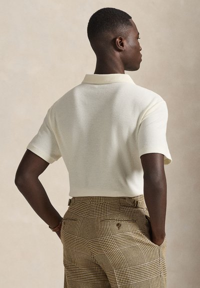 Άνδρας με κοντά μαλλιά, που φοράει υφασμάτινο λευκό κοντομάνικο πόλο και μπεζ καρό παντελόνι, με τα χέρια στις τσέπες, γυρισμένος προς τα πίσω.