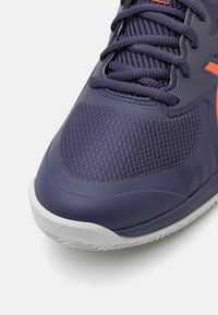 Pantof de sport violet, cu o combinație de plasă și cauciuc, având modele texturate, talpă albă și accente portocalii pe lateral.