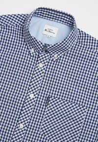 Camicia a quadri blu navy e bianca con colletto, taschino sul petto singolo e colletto interno azzurro chiaro. Presenta bottoni bianchi e etichetta.