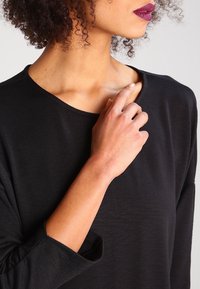 Haut noir à manches longues avec un col large, présentant une texture lisse. Une main touche délicatement le col.